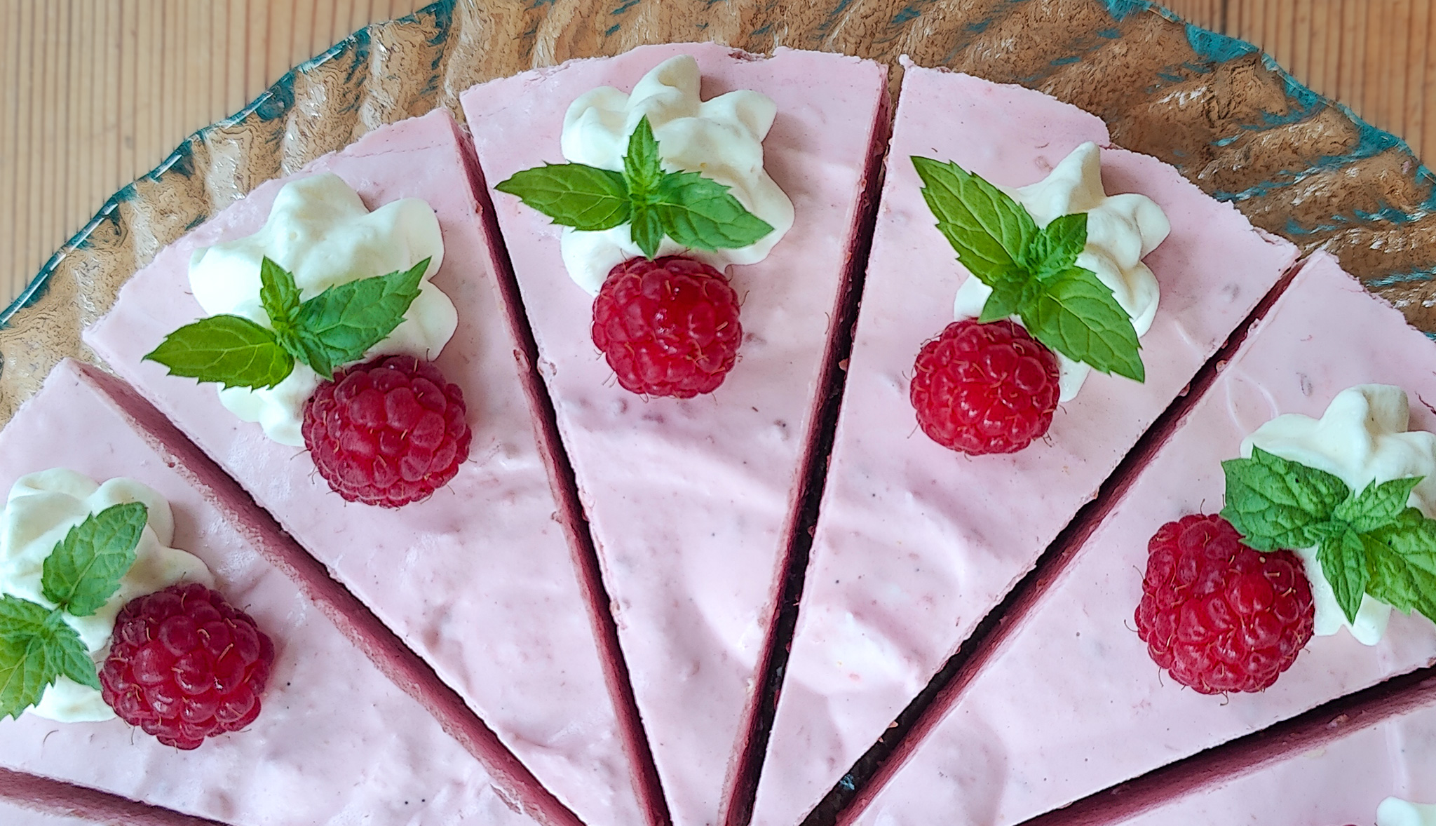 Ylhäältä kuvattu vadelmajuustokakku, jonka päällä kermapursotteita, isoja vadelmia ja mintunlehtiä.